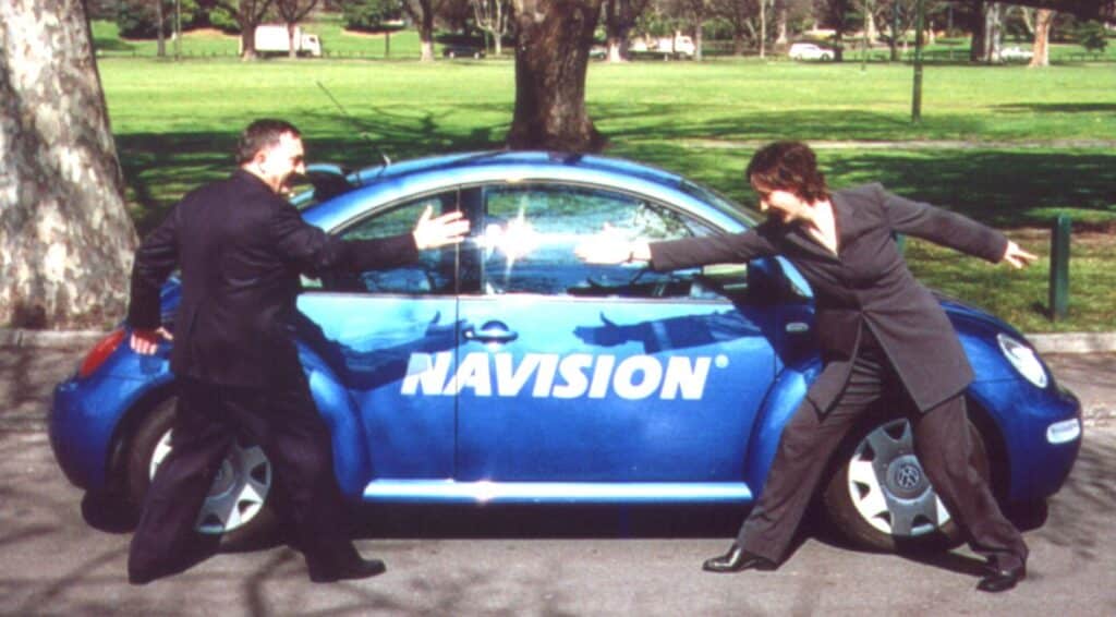 The side of a blue car with the word 
"Navision" across the door. Two people standing at either end of the car are posing in a running motion towards each other.